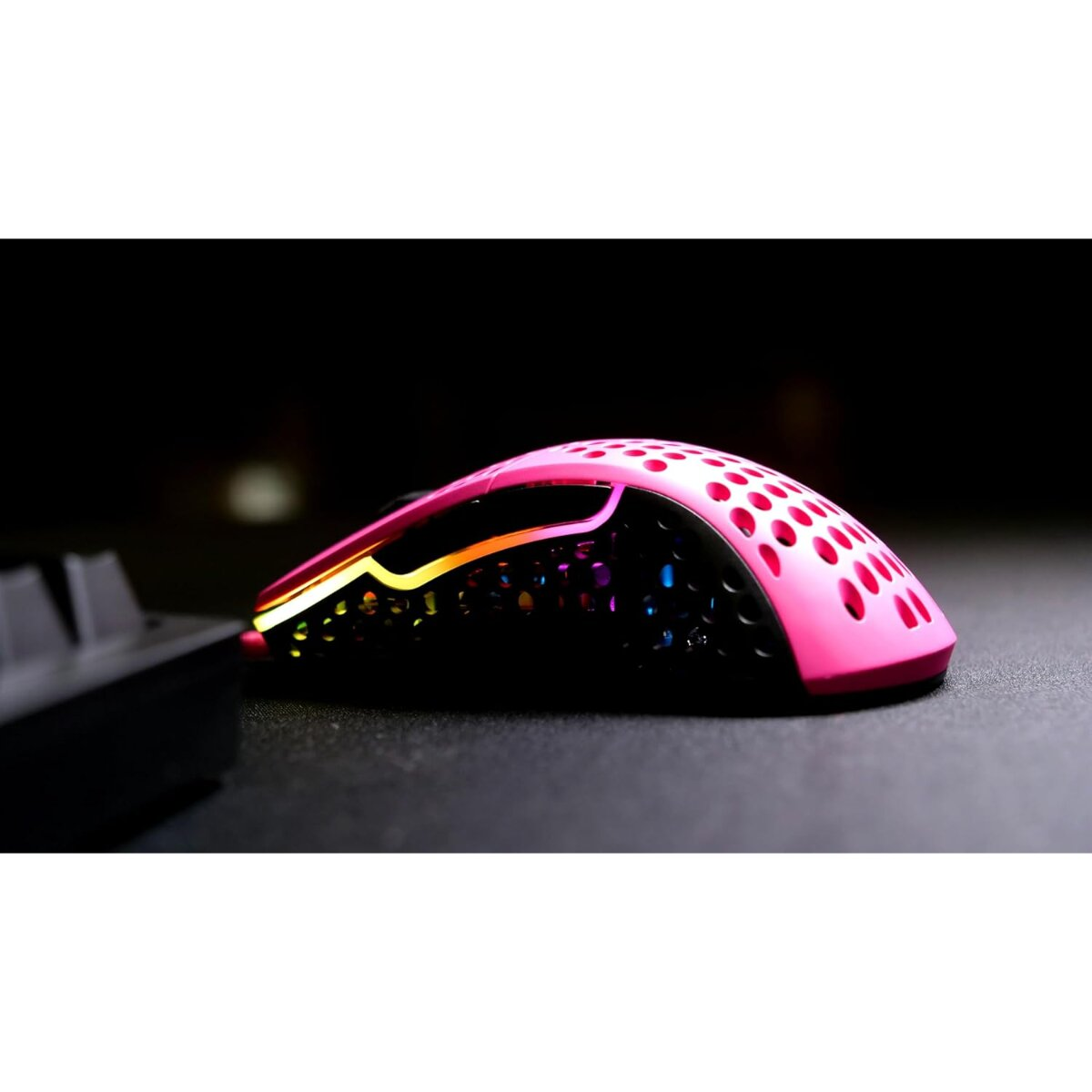 A pink and black gaming mouse with RGB lighting on a dark surface. The mouse has a honeycomb design and is angled to the left.