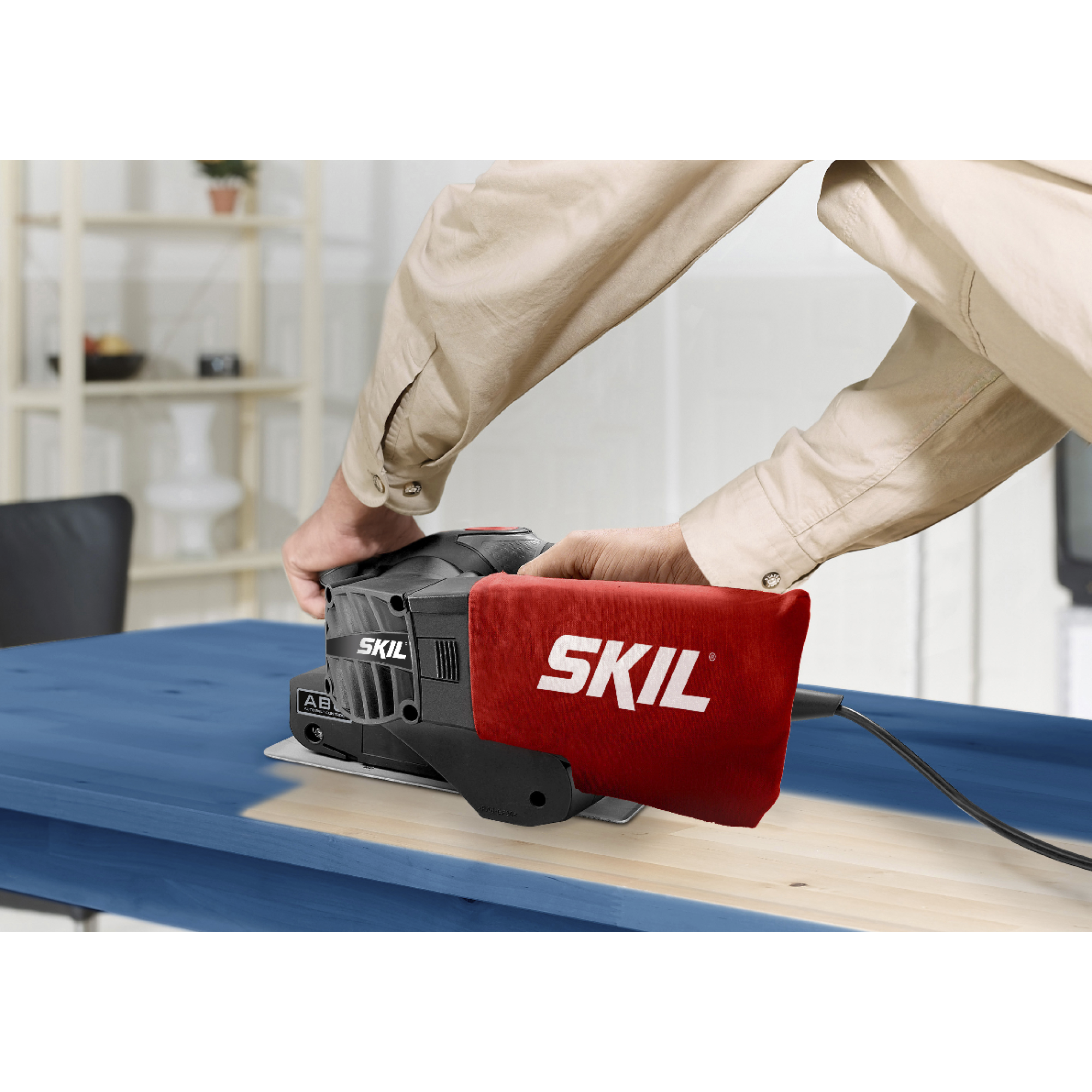 Een persoon schuurt een blauwe houten tafel met een SKIL schuurmachine. De schuurmachine is zwart met een rode stofzak.