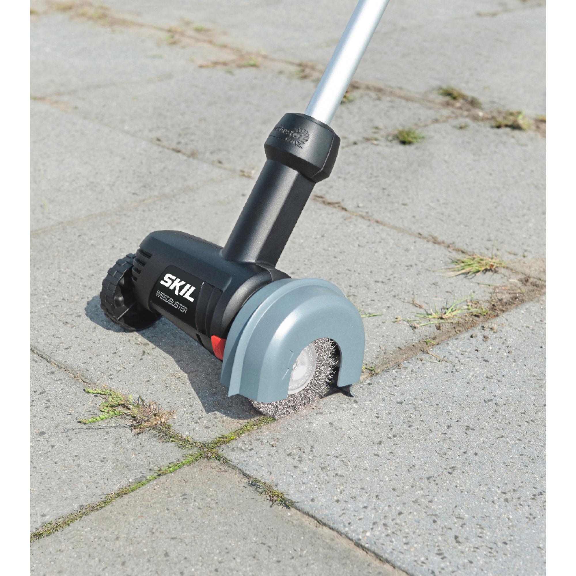 Een SKIL Weedeater staat op een grijze stenen ondergrond. Hij is zwart en grijs, met een draaiende borstel. Groen gras groeit in de scheuren.
