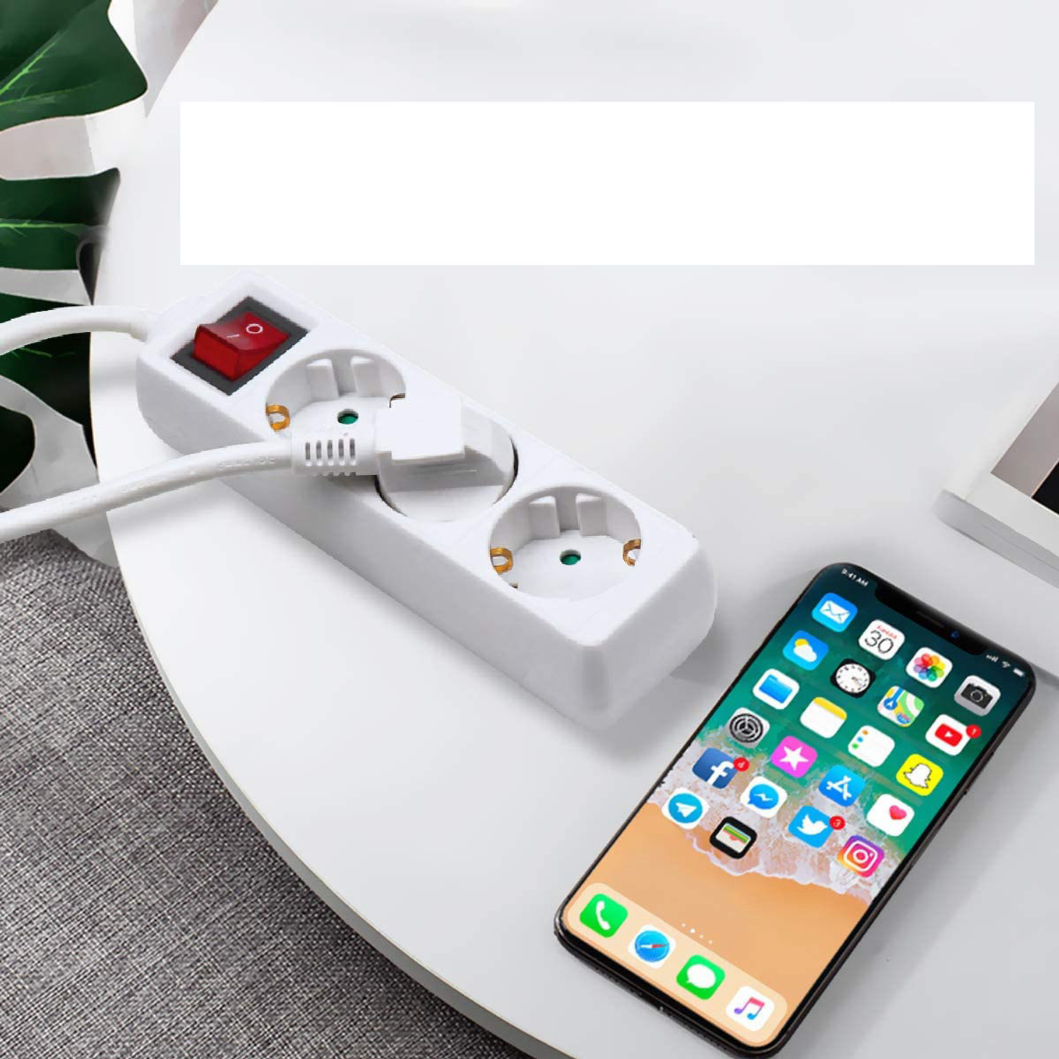 Ein weißes Verlängerungskabel mit rotem Schalter und ein Telefon mit App-Symbolen liegen auf einem weißen Tisch. Im Hintergrund ist eine Pflanze zu sehen.