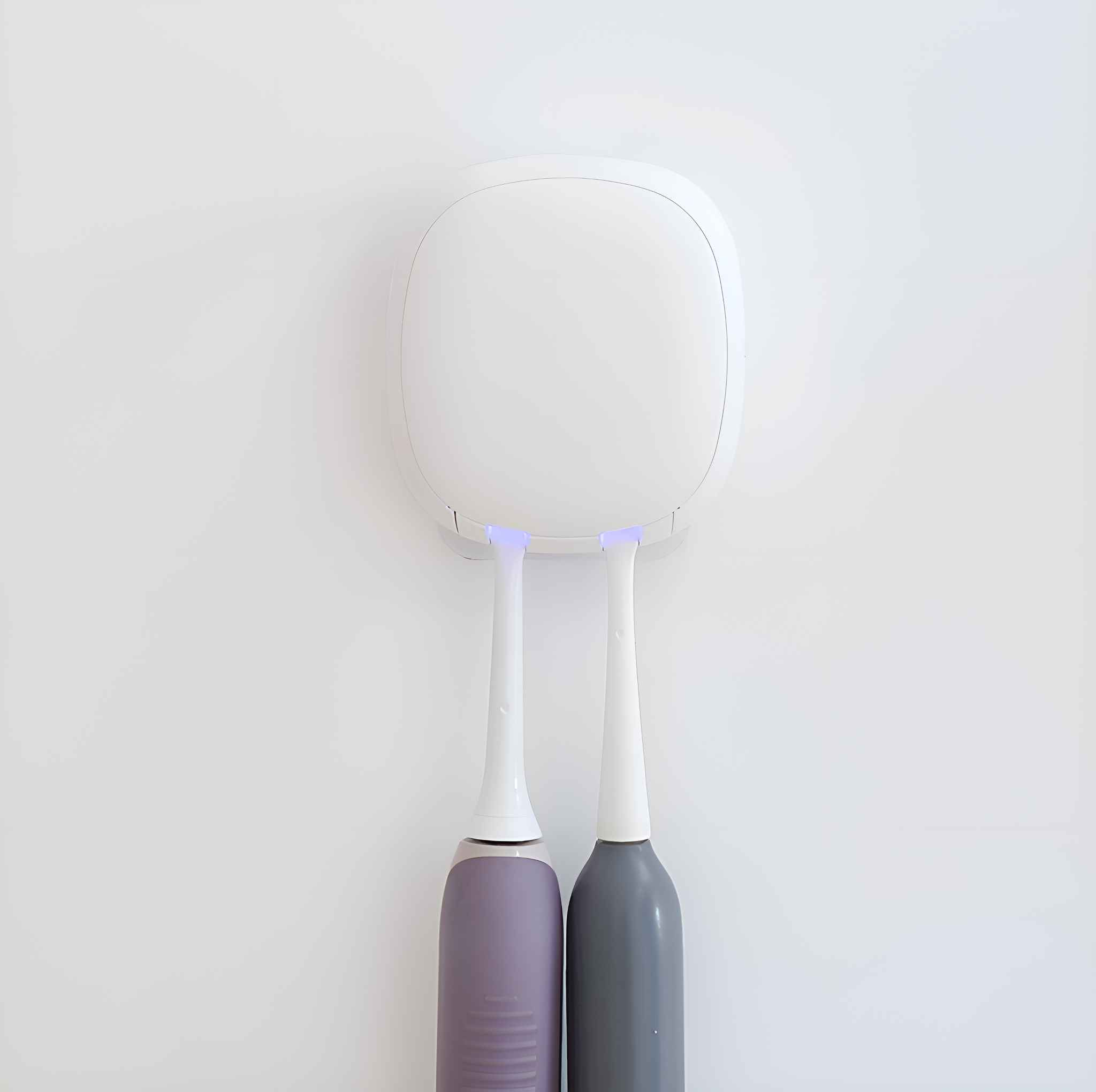Ein weißer Zahnbürstenhalter mit zwei elektrischen Zahnbürsten. Eine ist lila, die andere grau. Der Hintergrund ist eine weiße Wand.