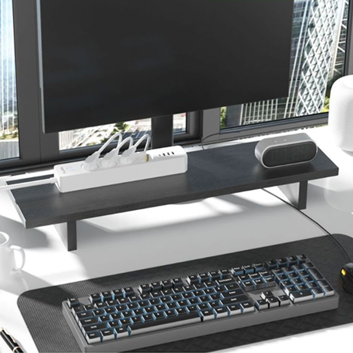Ein Schreibtisch-Setup mit Monitor, Tastatur, Steckdosenleiste und Uhr. Im Hintergrund eine Stadtlandschaft durch ein Fenster.