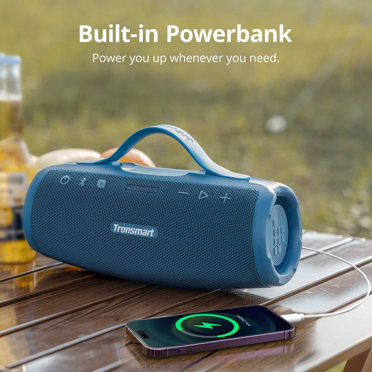 Ein blauer Tronsmart-Lautsprecher lädt ein Telefon auf einem Holztisch auf. Das Telefon zeigt 75 % geladen an. Im Hintergrund eine Wiese mit einer Flasche.