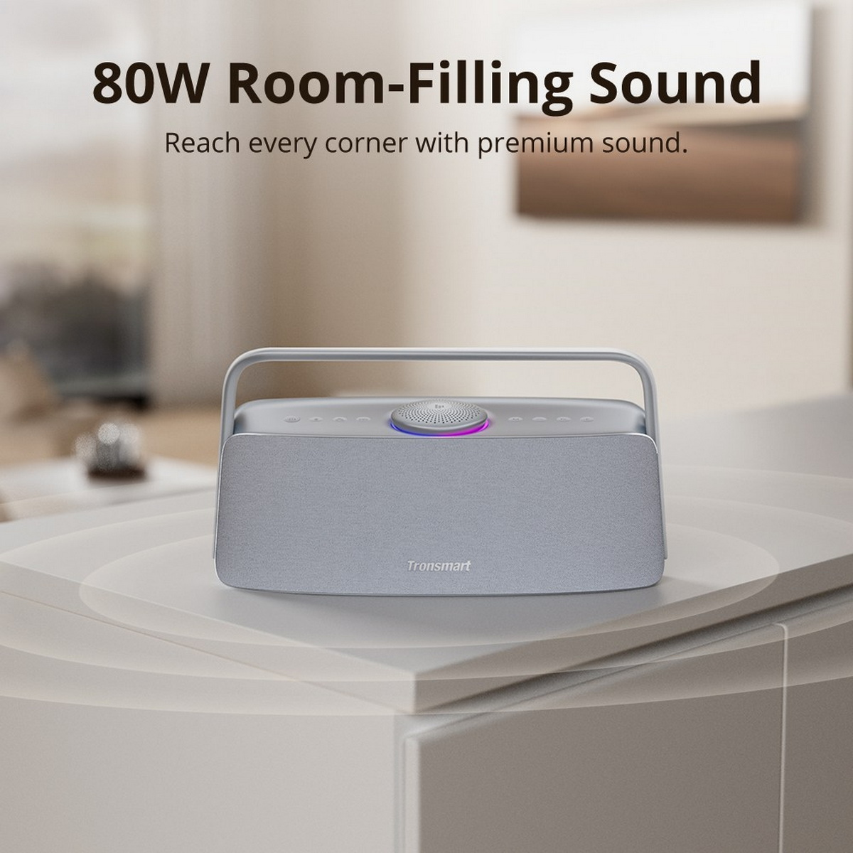Srebrny głośnik Tronsmart z uchwytem na białej powierzchni. Ma przyciski na górze i świecący środek. Tekst brzmi: '80W Room-Filling Sound'.