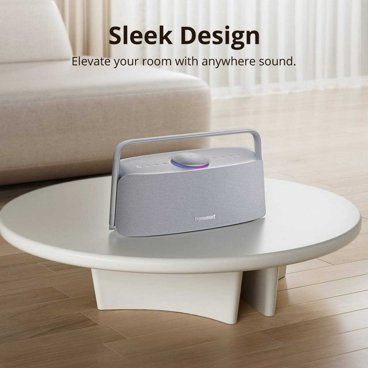 Szary głośnik z uchwytem stoi na białym stole. Kremowa sofa jest w tle. Tekst mówi 'Sleek Design'.