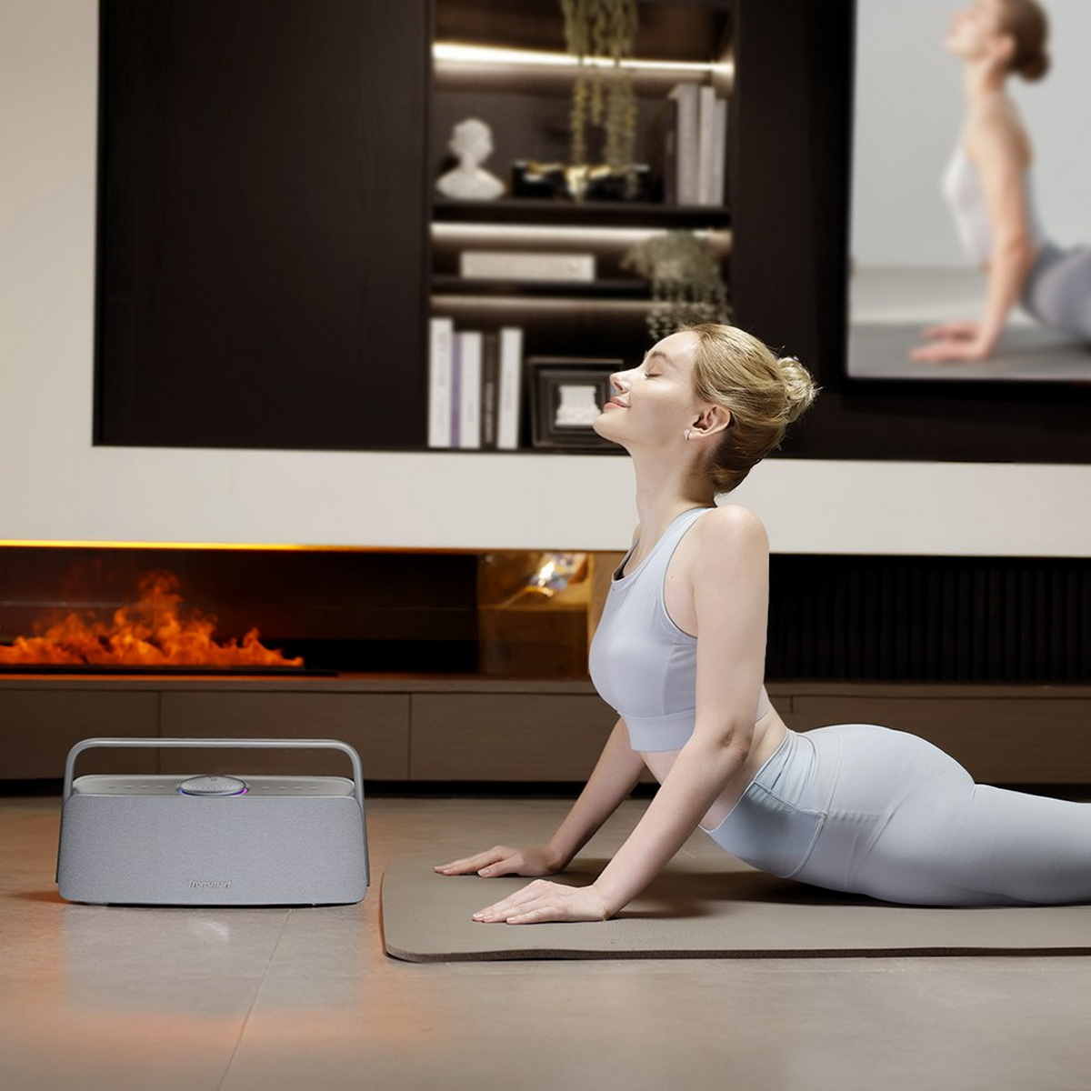 Eine Frau in Yoga-Pose, Augen geschlossen. Graue Matte, Lautsprecher und Kamin im Hintergrund. Ein Bildschirm zeigt die gleiche Pose.