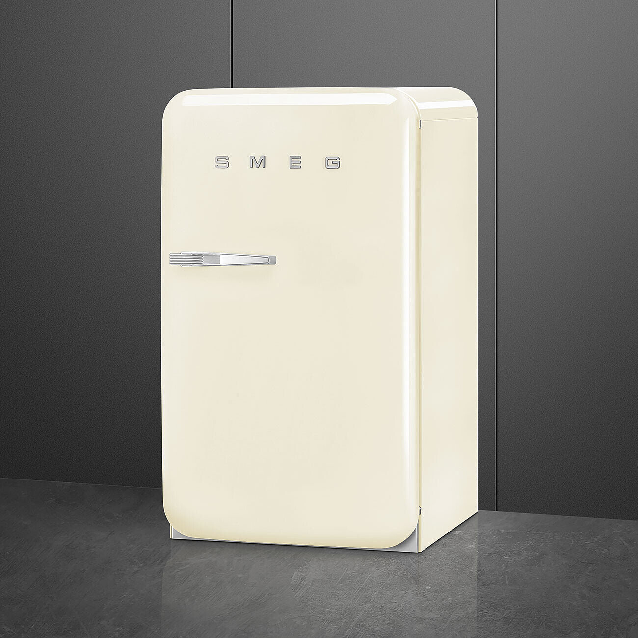 Un refrigerador SMEG de color crema está sobre una superficie oscura contra una pared gris oscuro. Tiene una manija y el nombre de la marca.