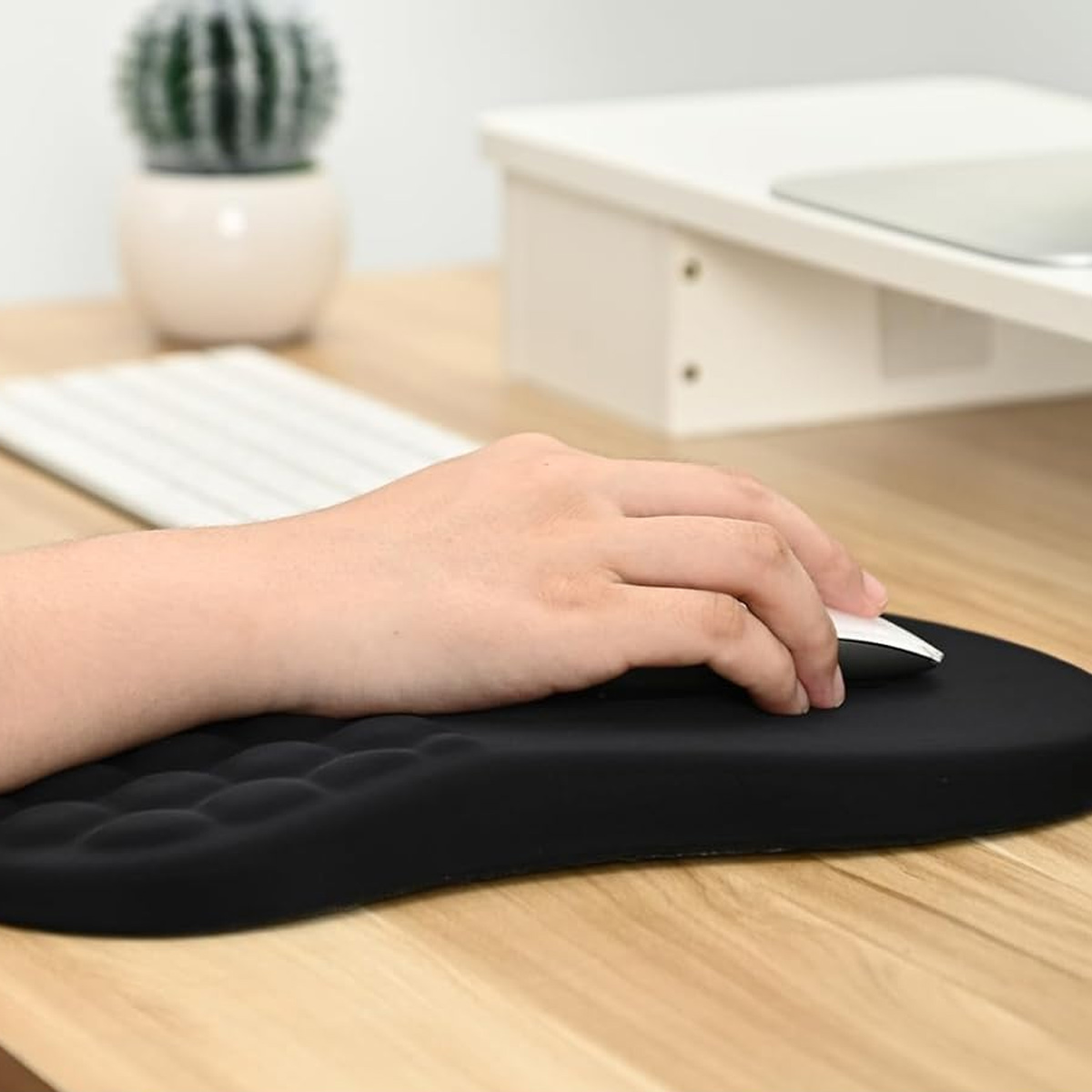 Eine Hand benutzt eine weiße Maus auf einem schwarzen Mauspapier. Eine Tastatur und ein Kaktus sind im Hintergrund auf einem Holztisch zu sehen.