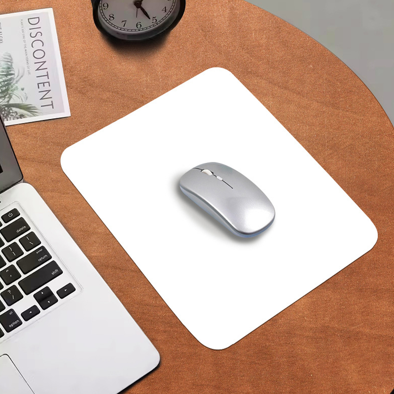 Eine silberne Computermaus auf einem weißen Mauspad auf einem braunen Tisch. Ein Laptop, eine Uhr und eine Zeitschrift sind ebenfalls vorhanden.