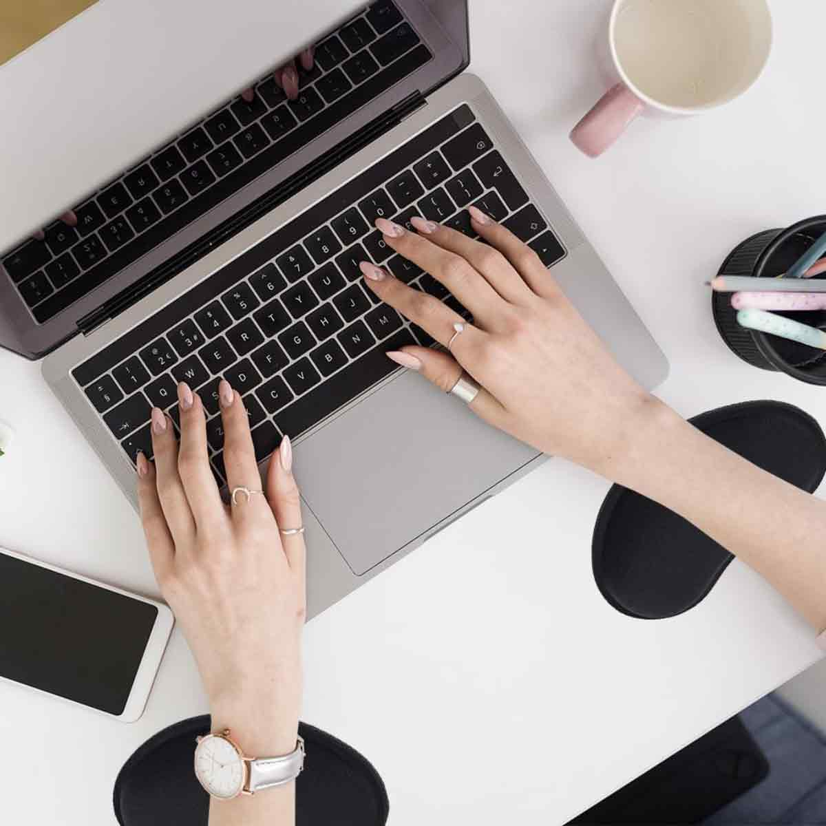 Hände tippen auf einer Laptop-Tastatur. Eine Uhr, Ringe, ein Telefon und eine Tasse stehen auf dem weißen Tisch. Schwarze Mauspads sind sichtbar.
