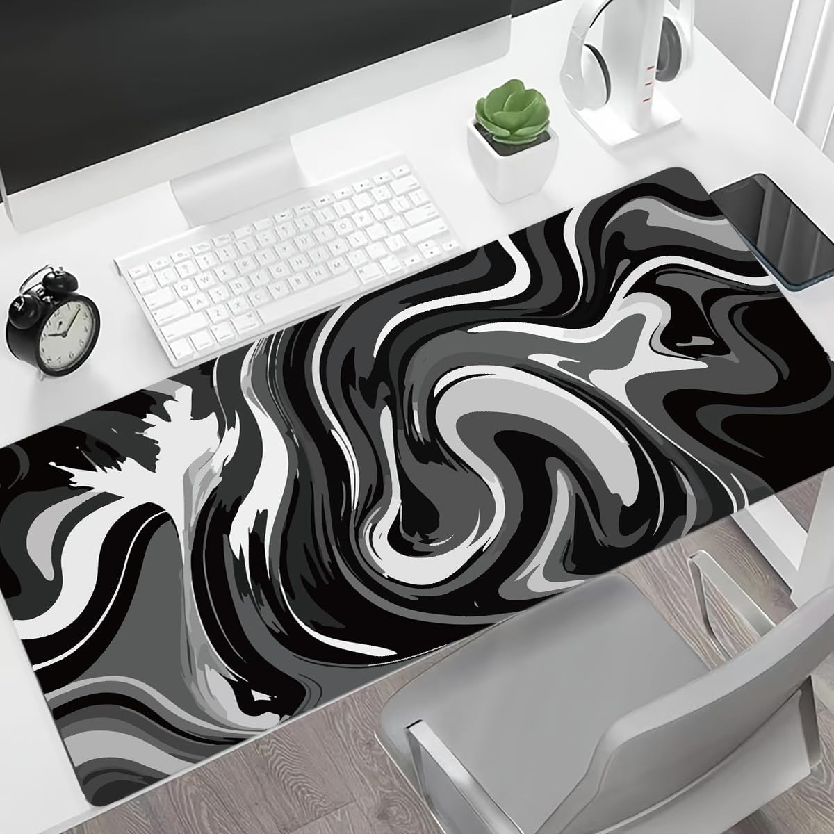 Ein Schreibtisch mit weißer Tastatur, Mauspad mit schwarz-weißem Wirbeldesign, Wecker, Kopfhörer und einer kleinen Pflanze.