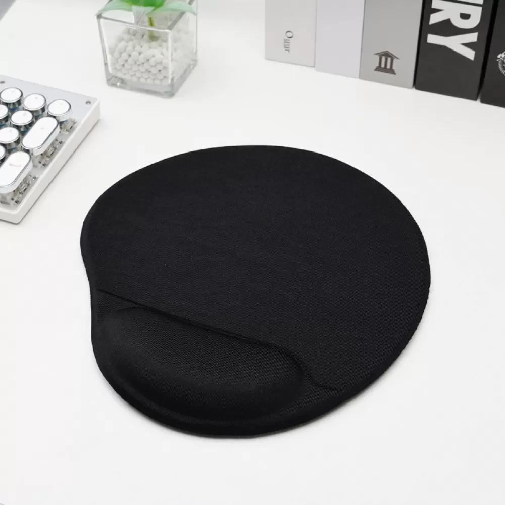Ein schwarzes Mauspads mit Handballenauflage liegt auf einer weißen Oberfläche. Tastatur und Bücher sind im Hintergrund, mit einer Pflanze.