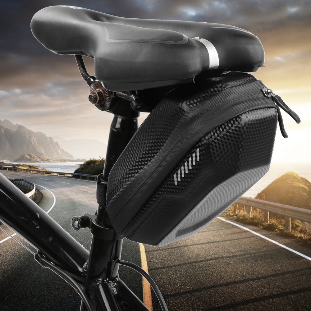 Schwarzer Fahrradsattel mit angebrachter Tasche. Im Hintergrund eine Küstenstraße mit Bergen unter einem bewölkten Himmel bei Sonnenuntergang.