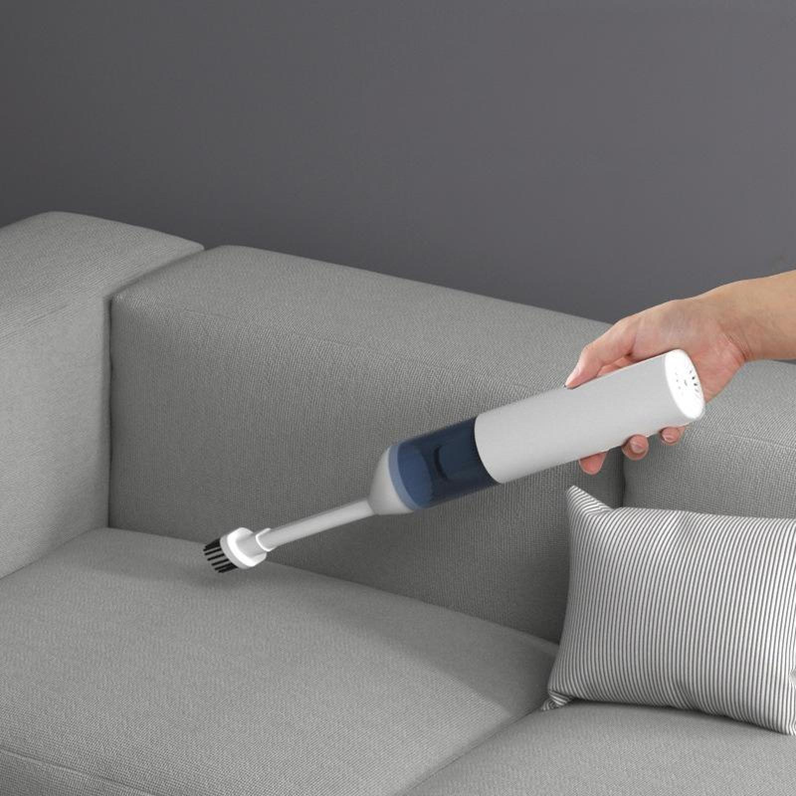 Eine Hand benutzt einen weiß-blauen Handstaubsauger, um ein graues Sofa zu reinigen. Ein gestreiftes Kissen liegt auf dem Sofa.
