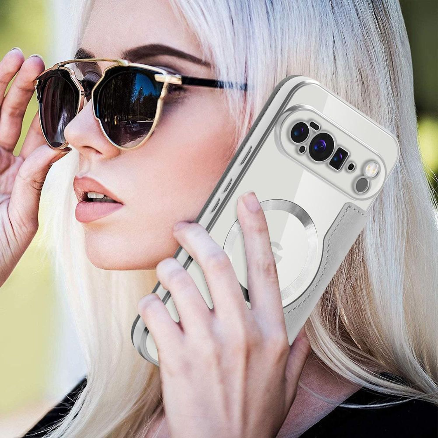 Blonde Frau mit Sonnenbrille, hält ein Smartphone in einer durchsichtigen Hülle. Sie ist draußen mit verschwommenem grünem Hintergrund.