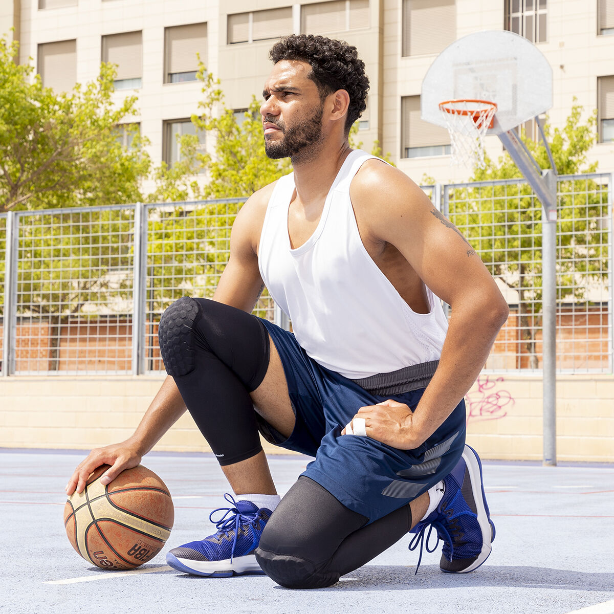Ein Mann kniet auf einem Basketballplatz und hält einen Ball. Er trägt ein weißes Tanktop, blaue Shorts, schwarze Knieschützer und blaue Schuhe.