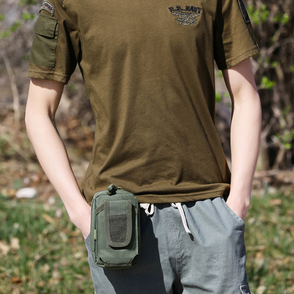 Eine Person in einem grünen Hemd und grauen Shorts mit einer grünen Tasche. Das Hemd hat ein US Navy-Logo. Hände in den Taschen.