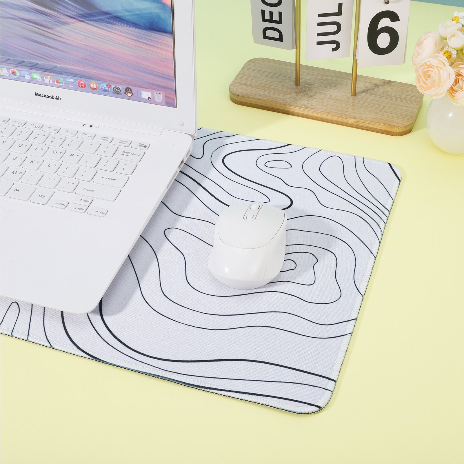 Ein weißer Laptop, eine Maus und ein Mauspapier mit topografischem Design liegen auf einer gelben Oberfläche. Ein Kalender und Blumen sind im Hintergrund.
