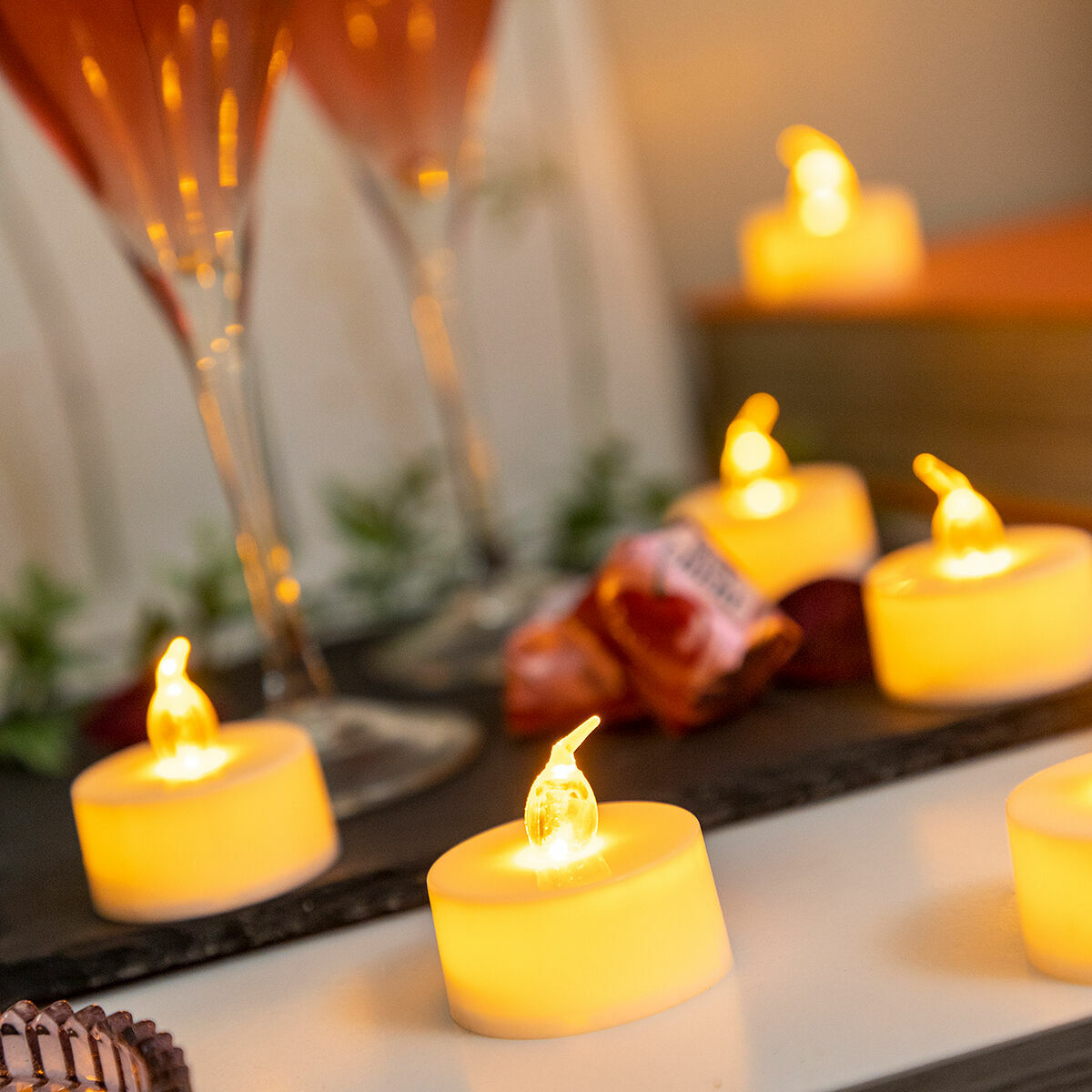 Primer plano de velas LED amarillas encendidas, dos copas de cóctel y un fondo borroso con golosinas. La escena es cálida y acogedora.