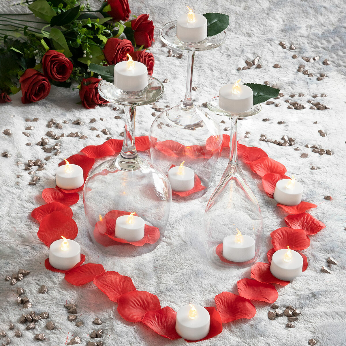 Escena romántica con velas en copas de vino, rodeada de pétalos de rosa. Rosas rojas en el fondo. Velas blancas encendidas.