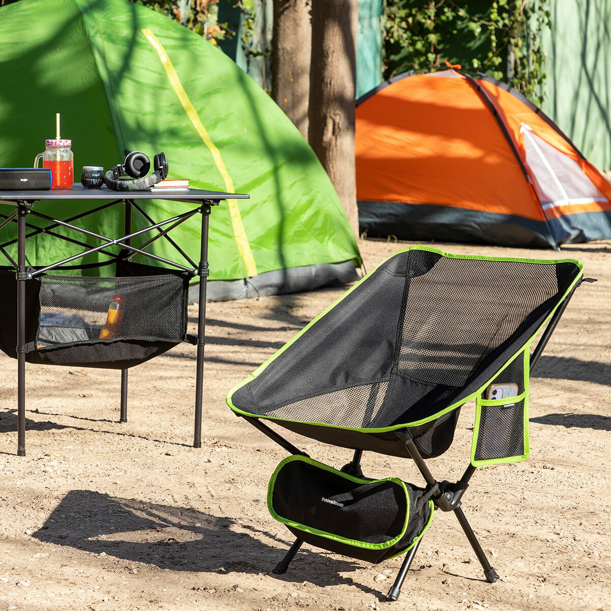 Eine Campingszene mit einem schwarz-grünen Stuhl im Vordergrund. Es gibt einen Tisch mit Getränken und Kopfhörern. Im Hintergrund stehen zwei Zelte.