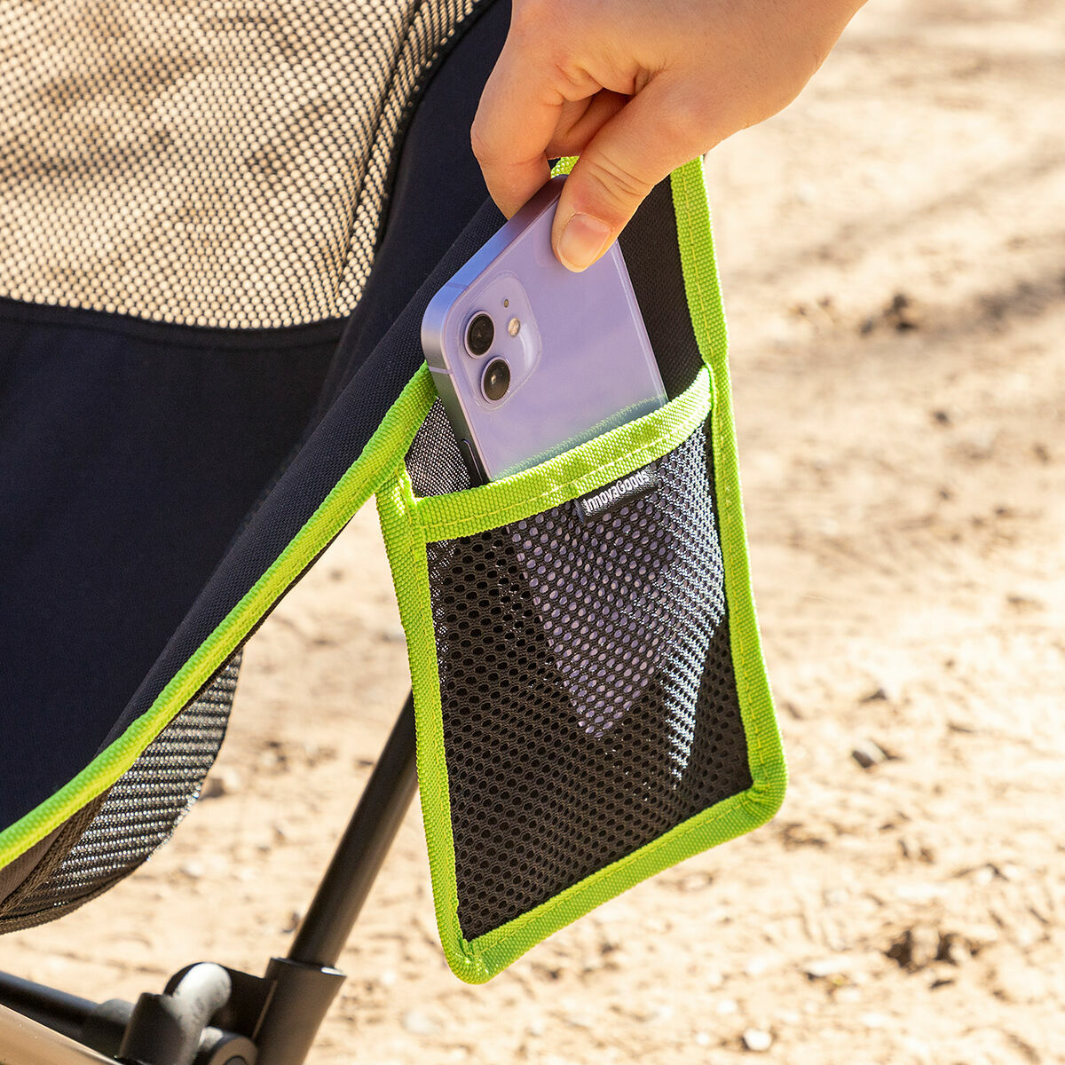 Eine Hand legt ein lila Telefon in eine schwarze Netztasche auf einem Klappstuhl. Die Tasche hat grüne Paspeln, und der Hintergrund ist Sand.
