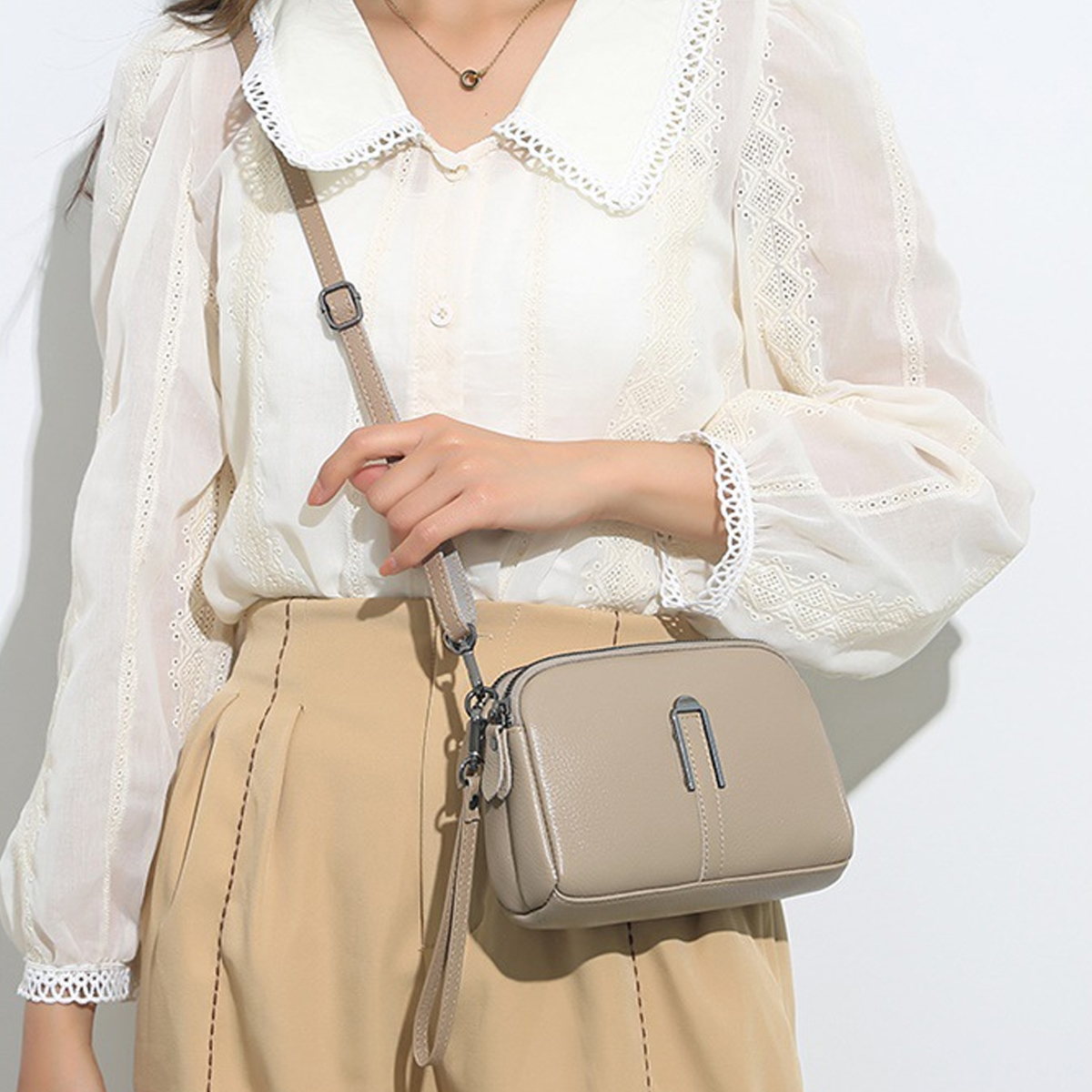 Eine Frau in einer weißen Bluse mit einer beigen Tasche. Die Tasche hat einen Schultergurt. Sie trägt eine Halskette und beige Hosen.