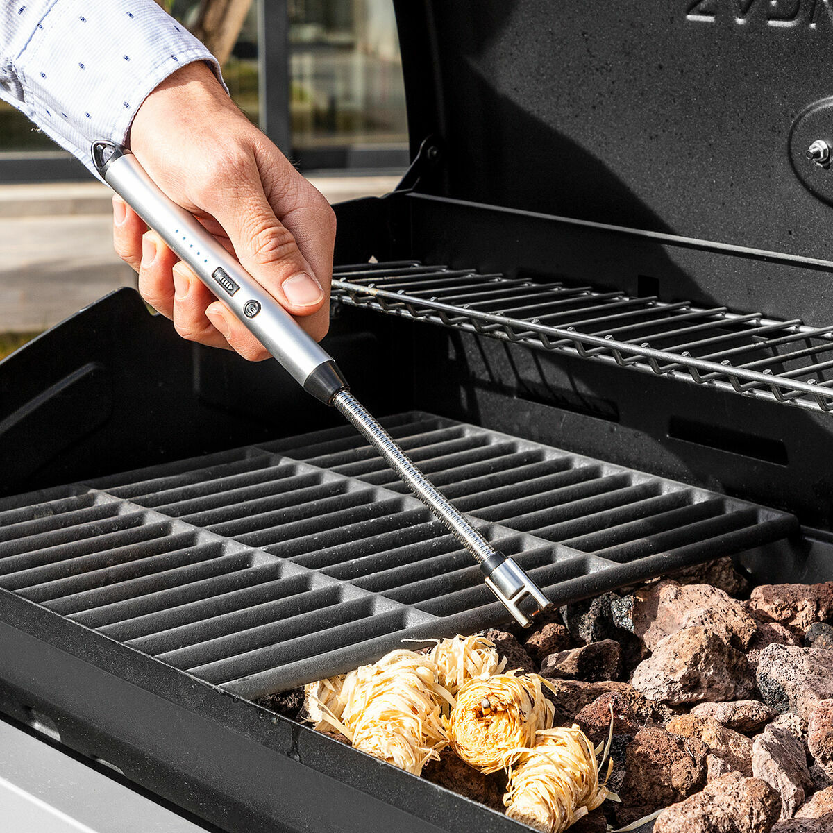 Eine Hand hält ein Feuerzeug über einem Grill mit Holzspänen und Kohlen. Der Grill ist offen, und eine Hand hält das Feuerzeug.
