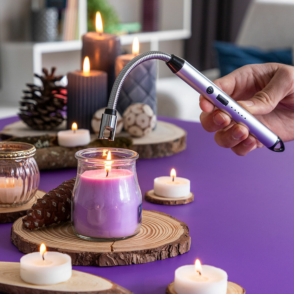 Eine Hand hält ein Feuerzeug über einer brennenden Kerze. Auf einer lila Oberfläche befinden sich mehrere andere Kerzen und Dekorationsgegenstände.