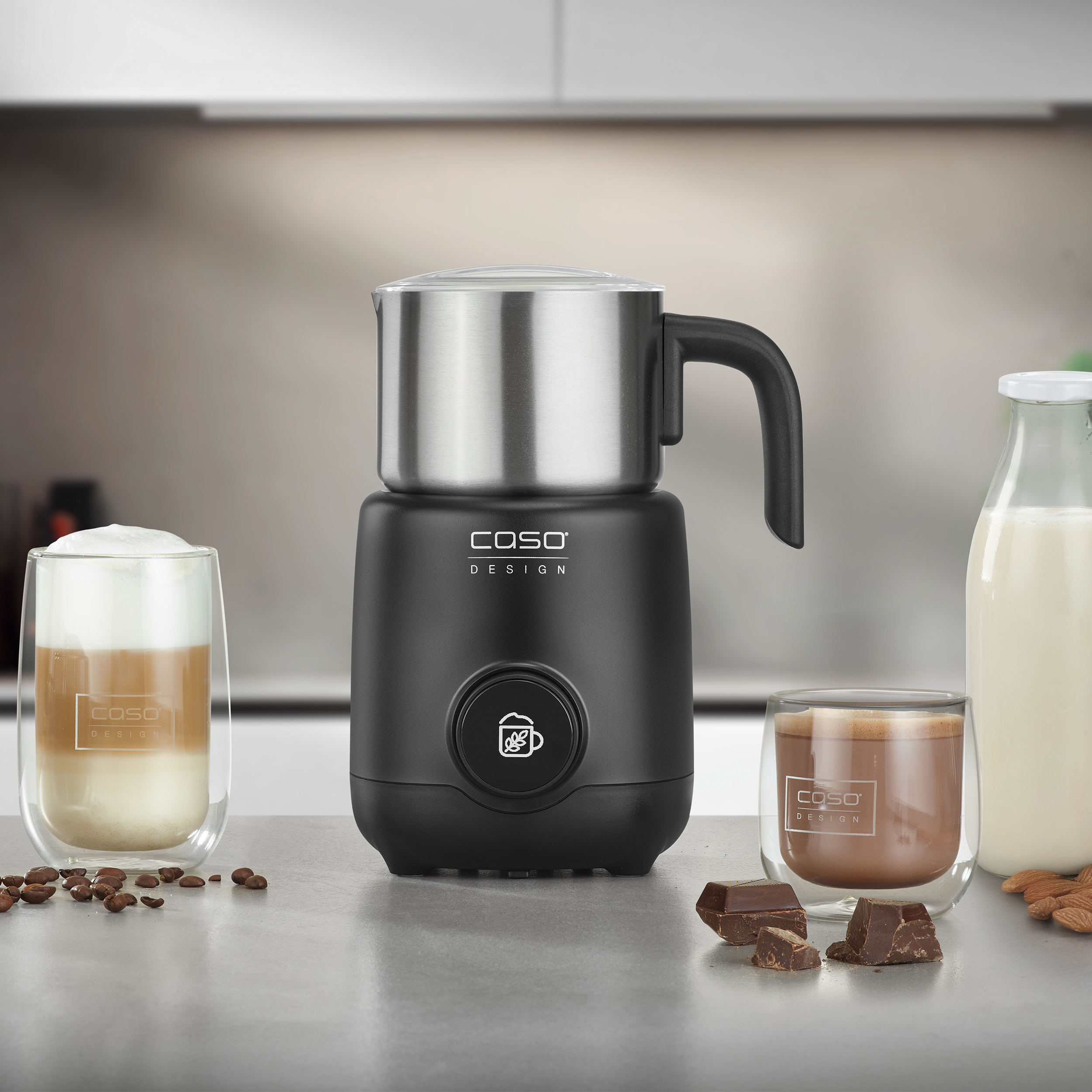 Ein schwarzer und silberner Milchaufschäumer mit zwei Kaffeegläsern, einer Milchflasche und Schokoladenstücken auf einer grauen Theke.