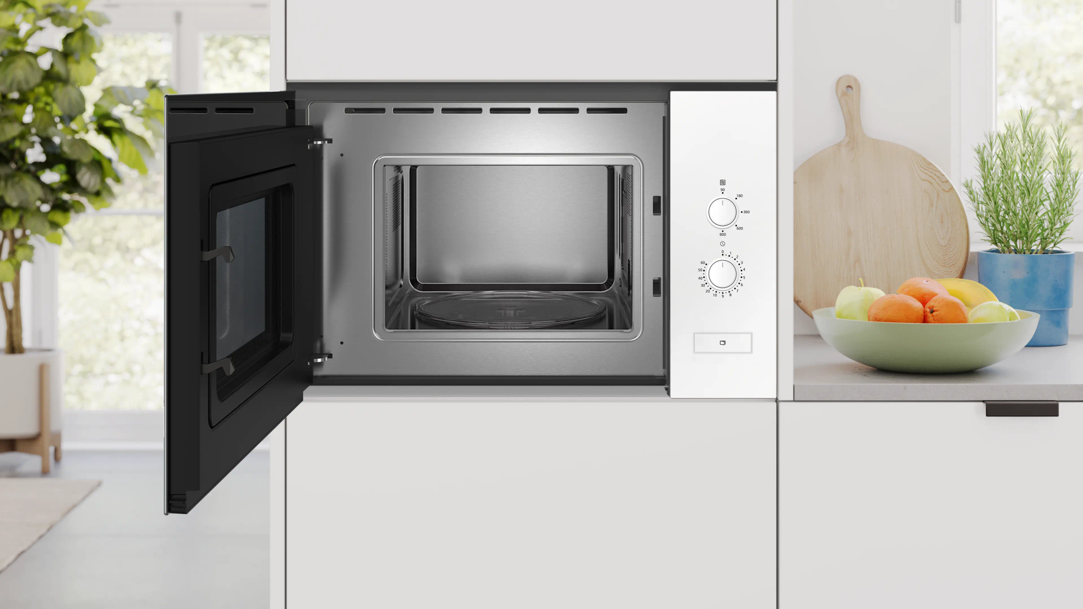 Otwarta kuchenka mikrofalowa w białej kuchni. Miska z owocami obok deski do krojenia. Roślina w tle.