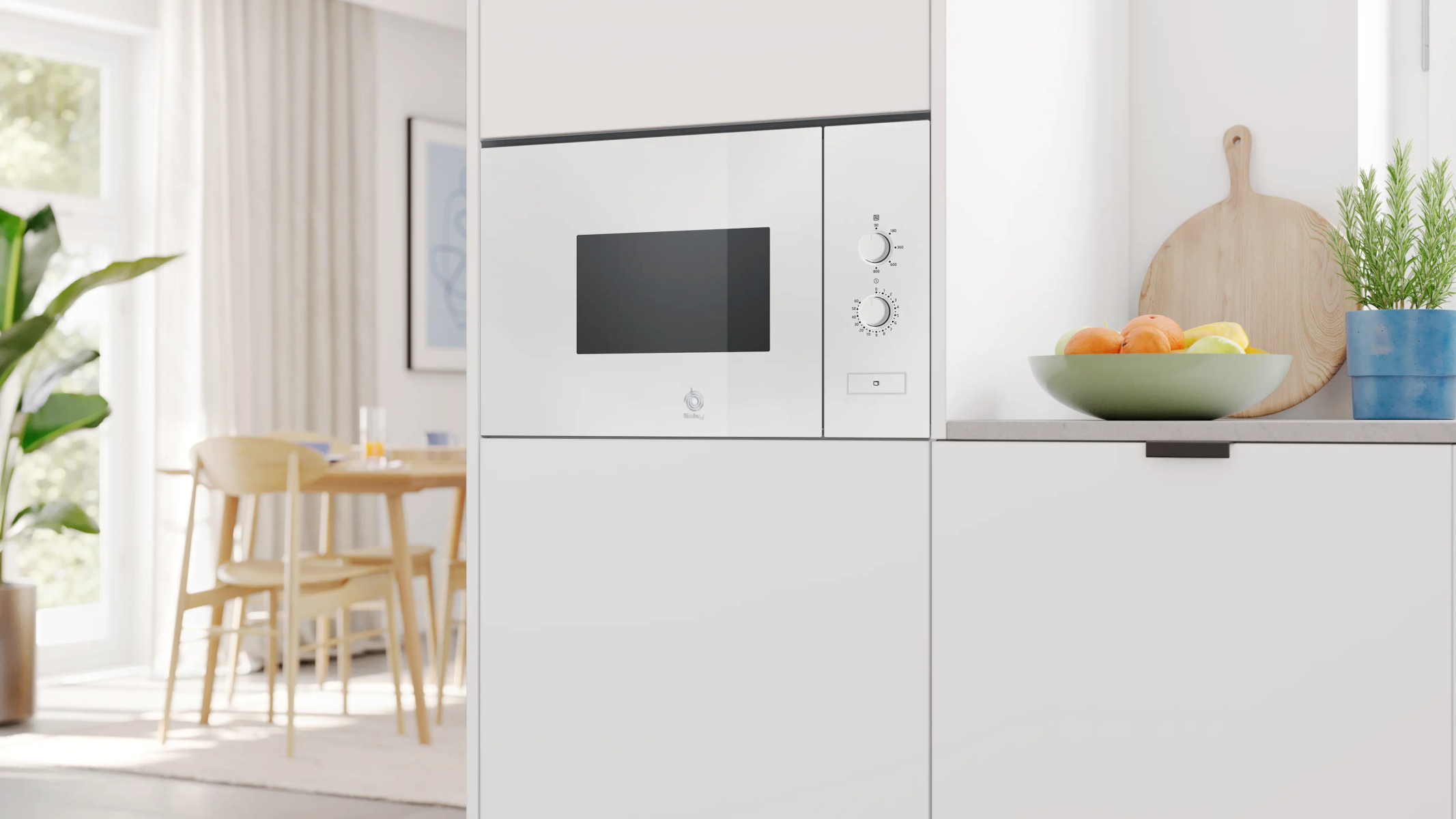 Kuchnia z białą kuchenką mikrofalową, owocami w misce i rośliną. W tle widoczny jest stół jadalniany.