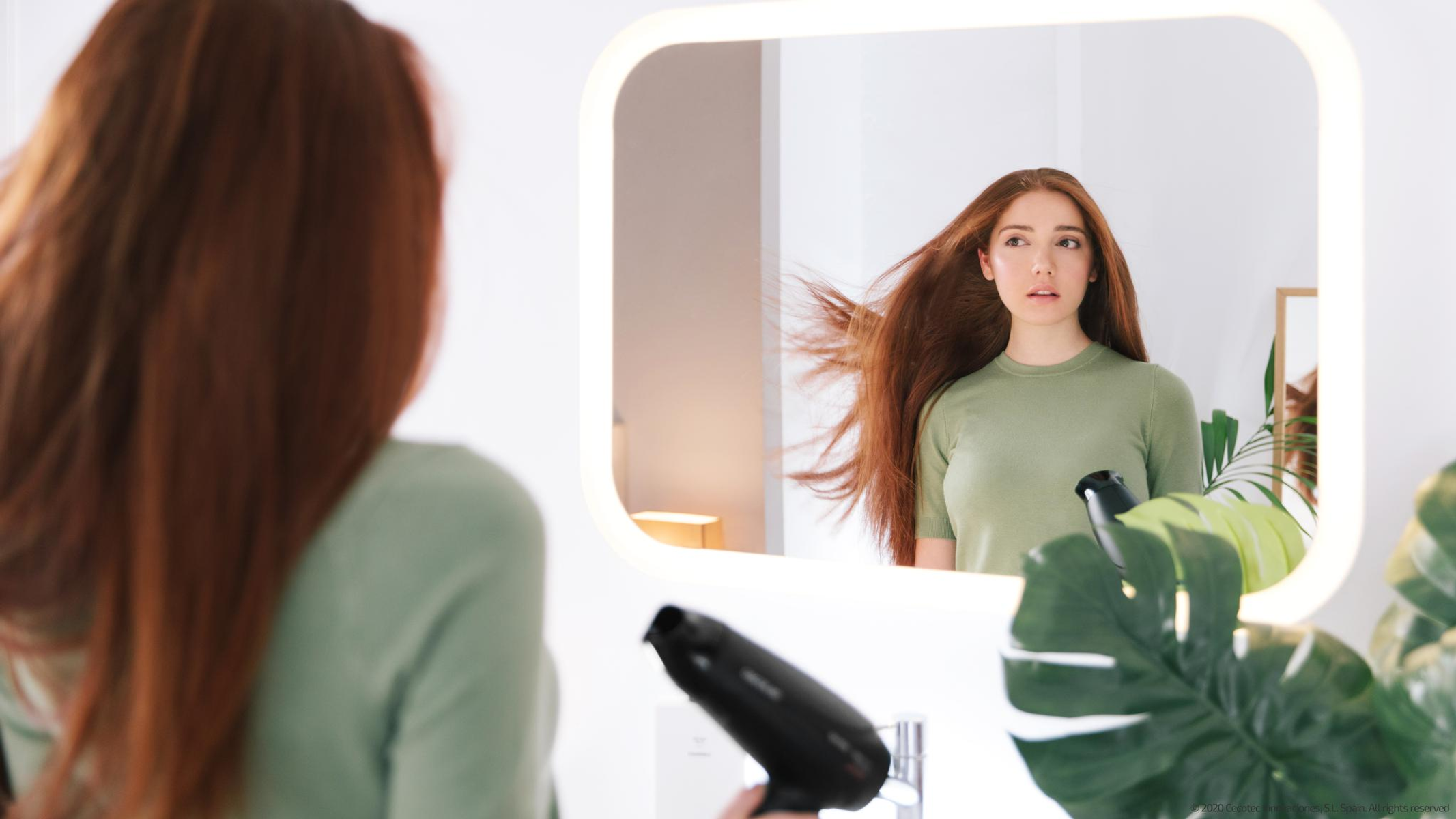 Una mujer se mira en un espejo mientras se seca el pelo. Sostiene un secador de pelo. El espejo refleja su pelo al viento.