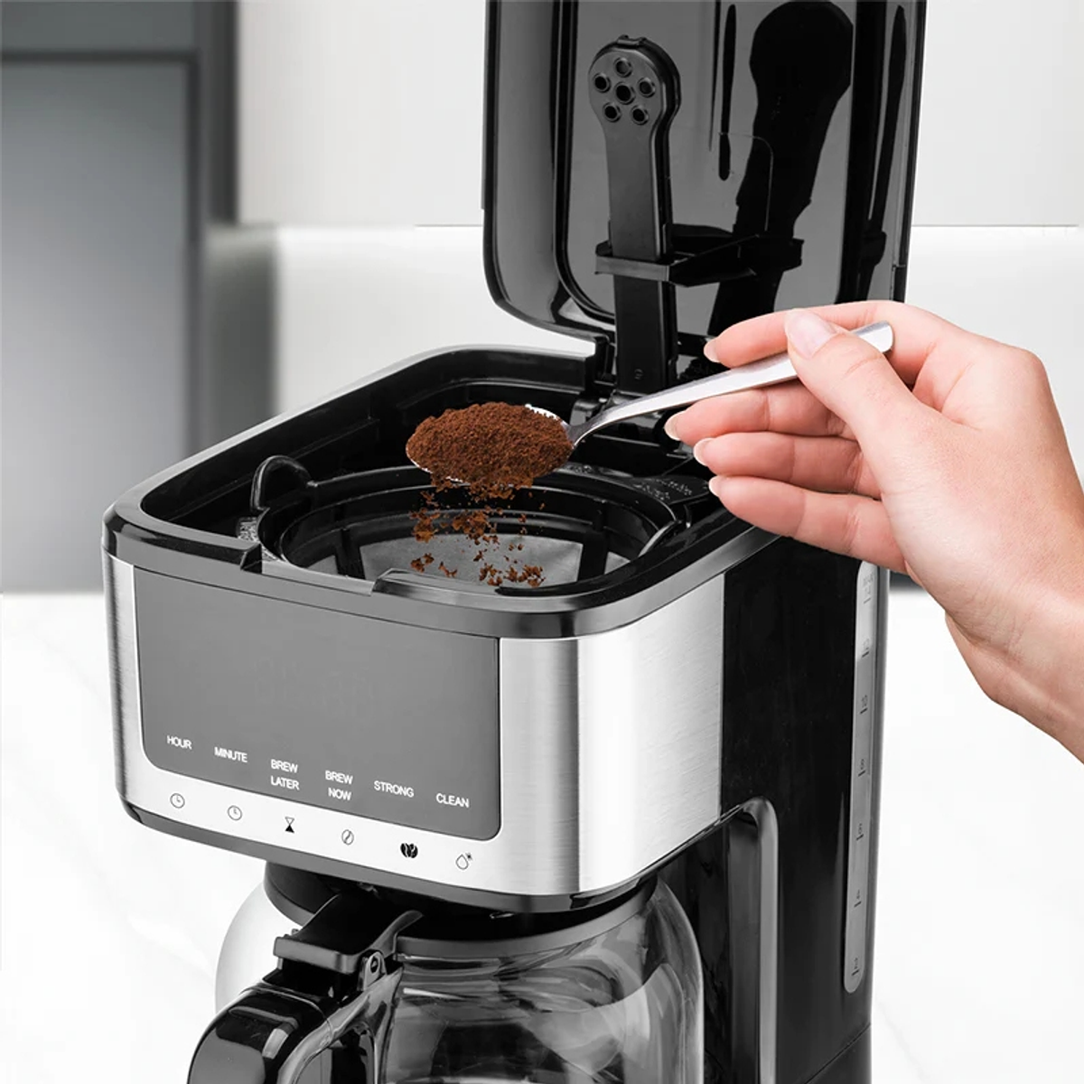Una persona versa fondi di caffè in una macchina per il caffè con un cucchiaio. La macchina è nera e argento, lo sfondo è bianco.
