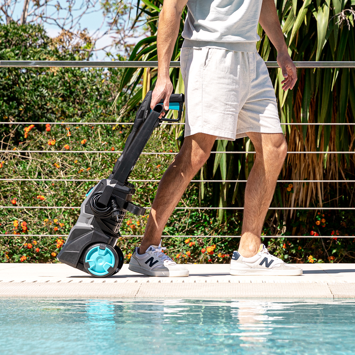 Una persona cammina con un'idropulitrice vicino a una piscina. La lavatrice è nera e turchese. La persona indossa pantaloncini e scarpe da ginnastica.