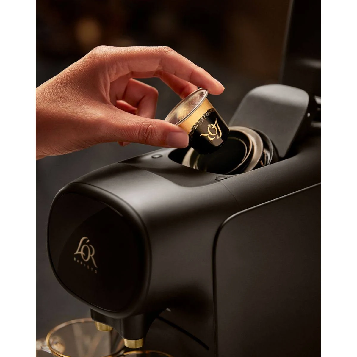 Una mano colocando una cápsula de café en una máquina L'Or Barista negra. La cápsula es negra y dorada. Fondo oscuro.