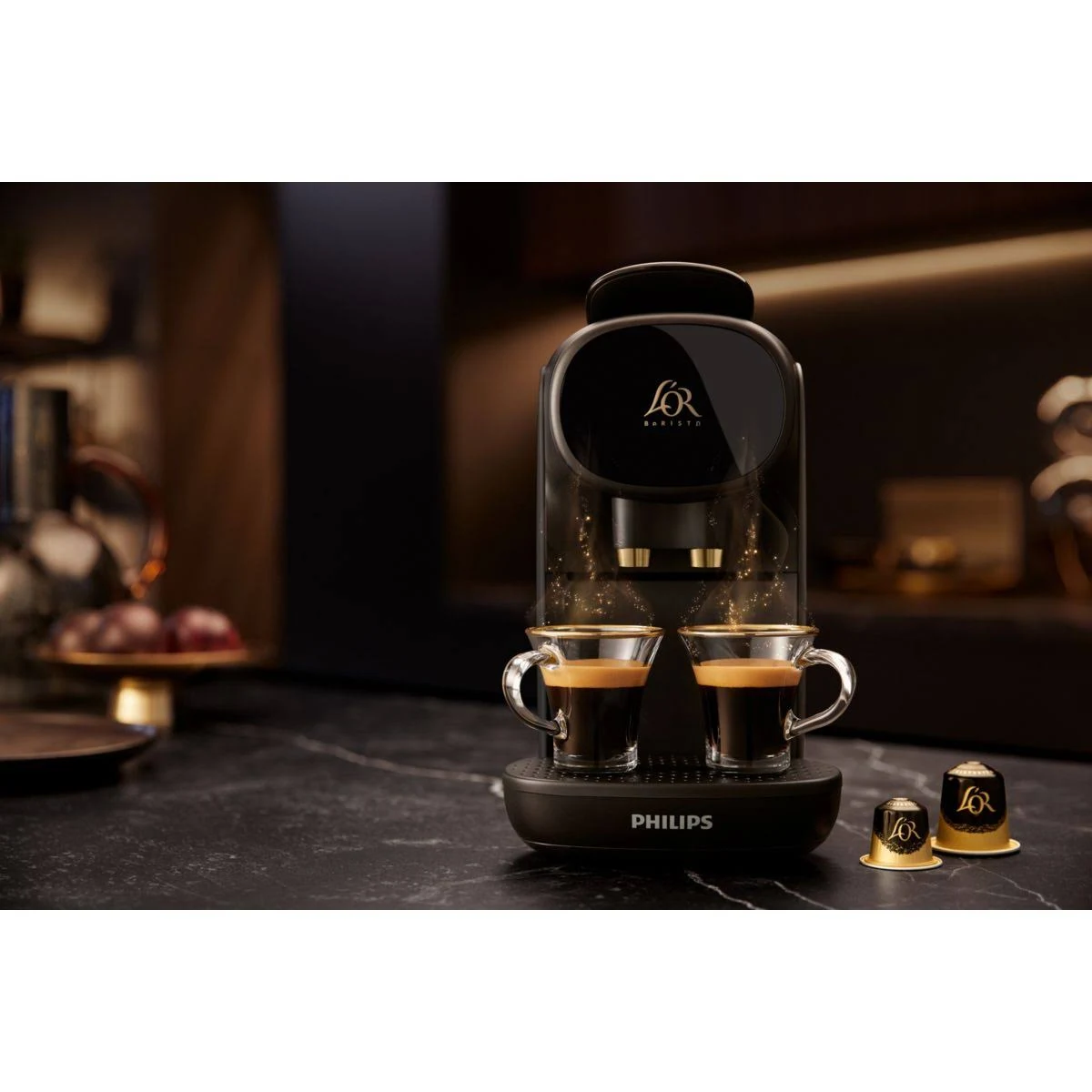 Una cafetera L'OR Barista negra y dorada con dos tazas de espresso. Dos cápsulas de café a la derecha.