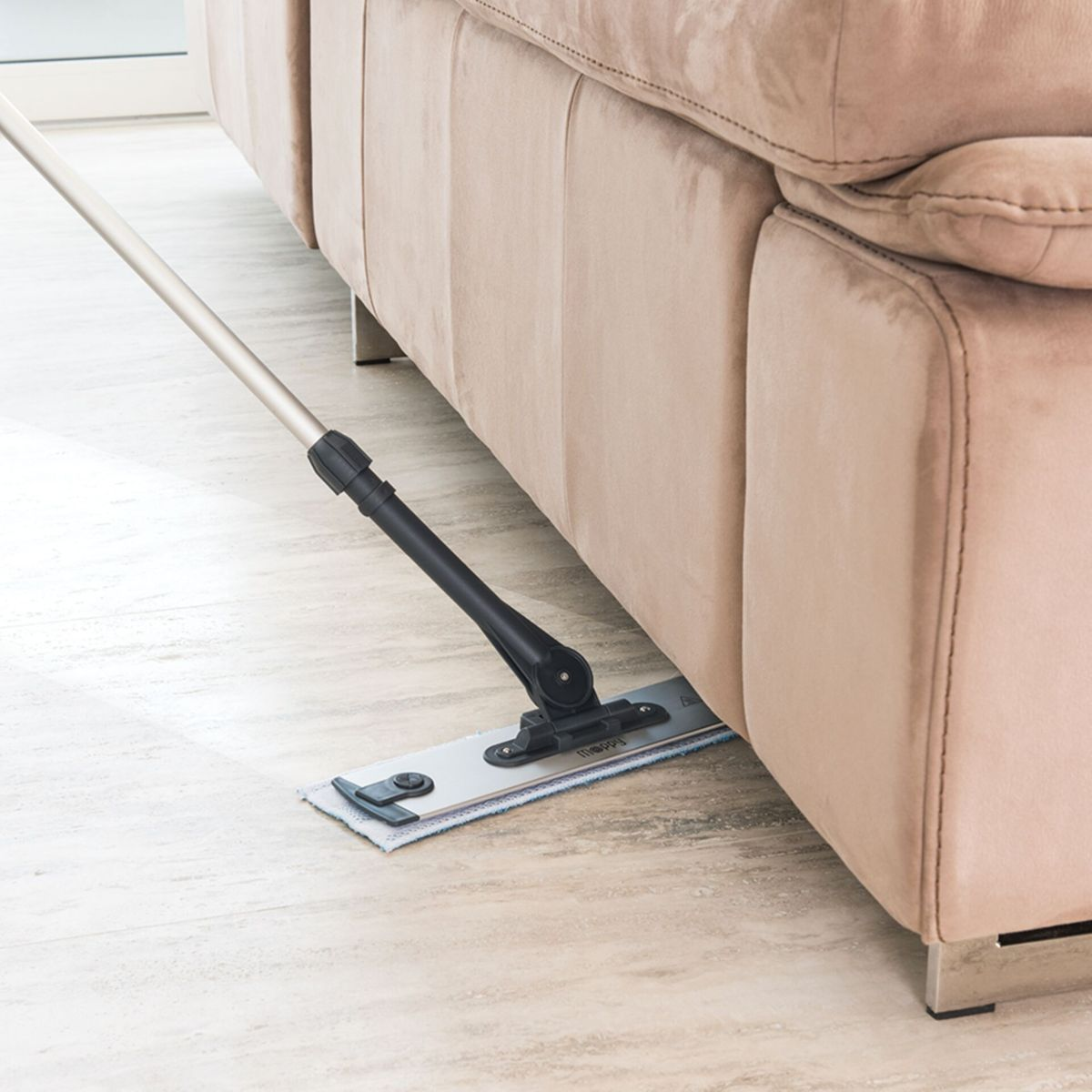 Mop czyści pod beżową sofą. Mop ma srebrny uchwyt i szarą nakładkę czyszczącą. Podłoga jest jasna, wyłożona płytkami.