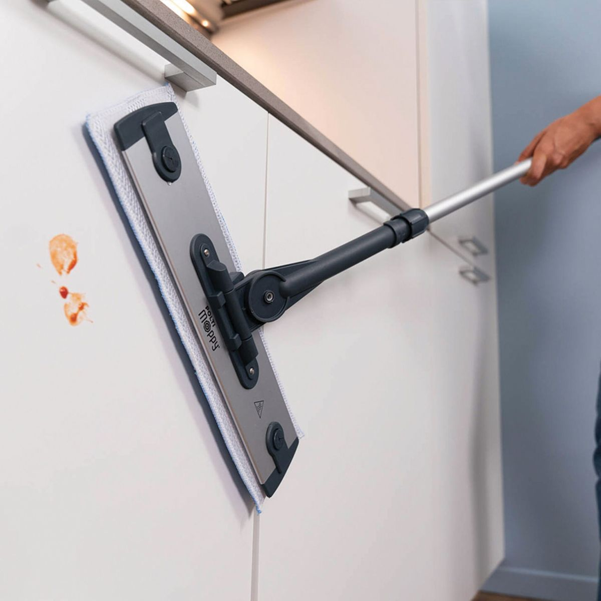 Osoba używa mopa do czyszczenia białej szafki kuchennej z plamami po sosie pomidorowym. Mop jest srebrny i czarny.