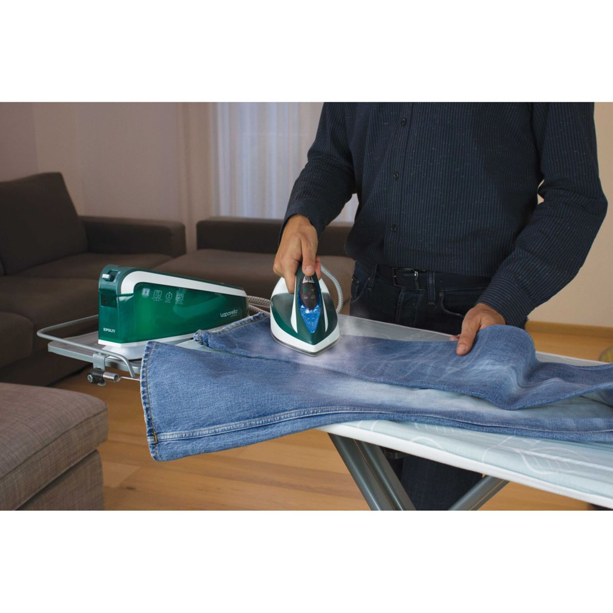 Eine Person bügelt eine blaue Jeans auf einem Bügelbrett. Ein grün-weißes Bügeleisen steht neben dem Brett. Im Hintergrund ist eine Couch zu sehen.
