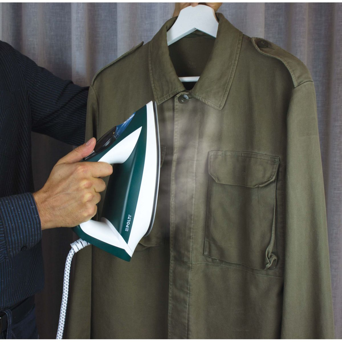 Eine Person bügelt eine olivgrüne Jacke mit einem grün-weißen Bügeleisen. Dampf steigt aus dem Bügeleisen auf. Die Jacke hängt an einem weißen Kleiderbügel.