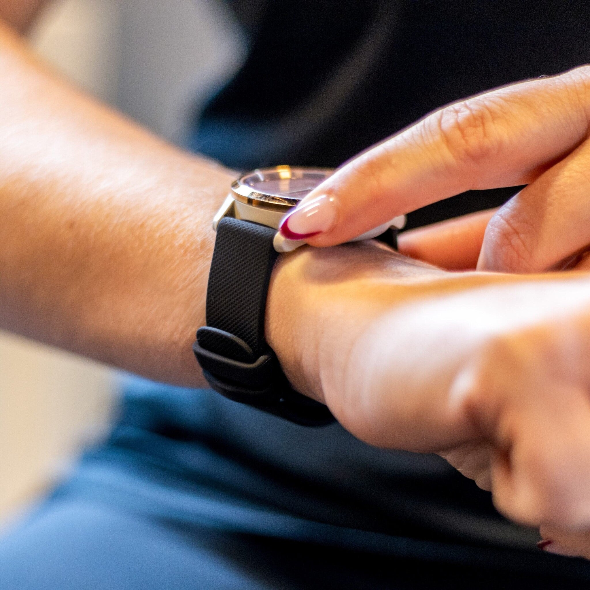 Eine Hand berührt eine Smartwatch. Die Uhr hat ein schwarzes Armband und eine goldene Lünette. Die Nägel der Person sind lackiert.