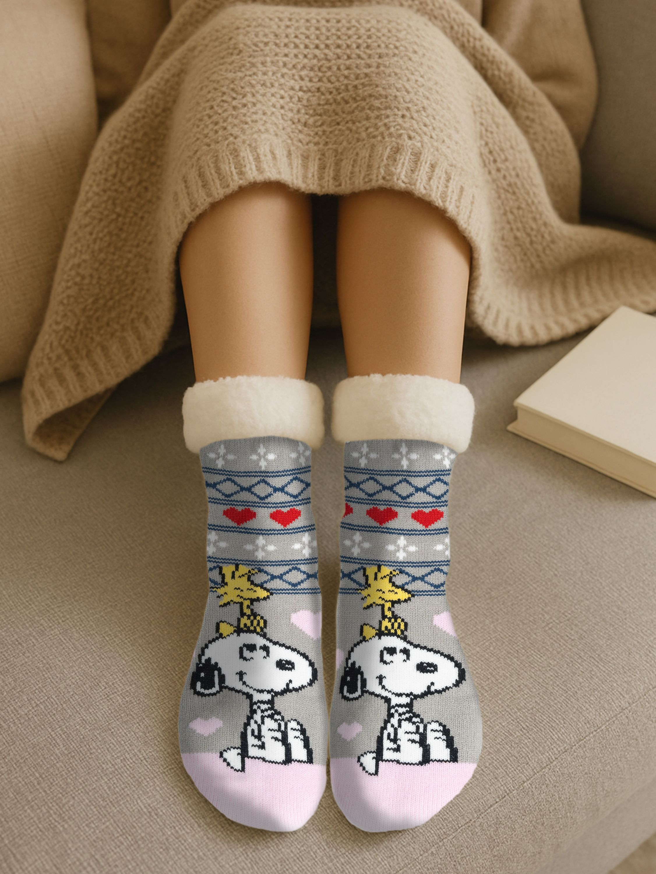 Person trägt Snoopy-Socken, sitzt auf einem Sofa. Die Socken sind grau, mit Snoopy, Herzen und gelbem Woodstock. Ein Buch ist in der Nähe.