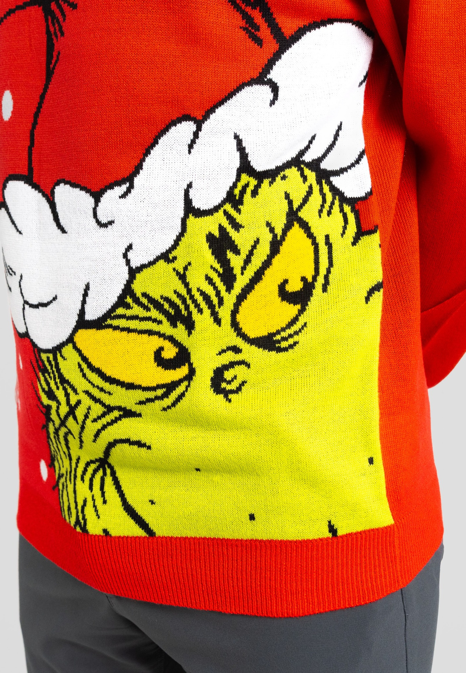 Eine Person trägt einen roten Pullover mit dem Gesicht des Grinch. Der Grinch hat gelbe Haut und weiße Haare. Graue Hose.