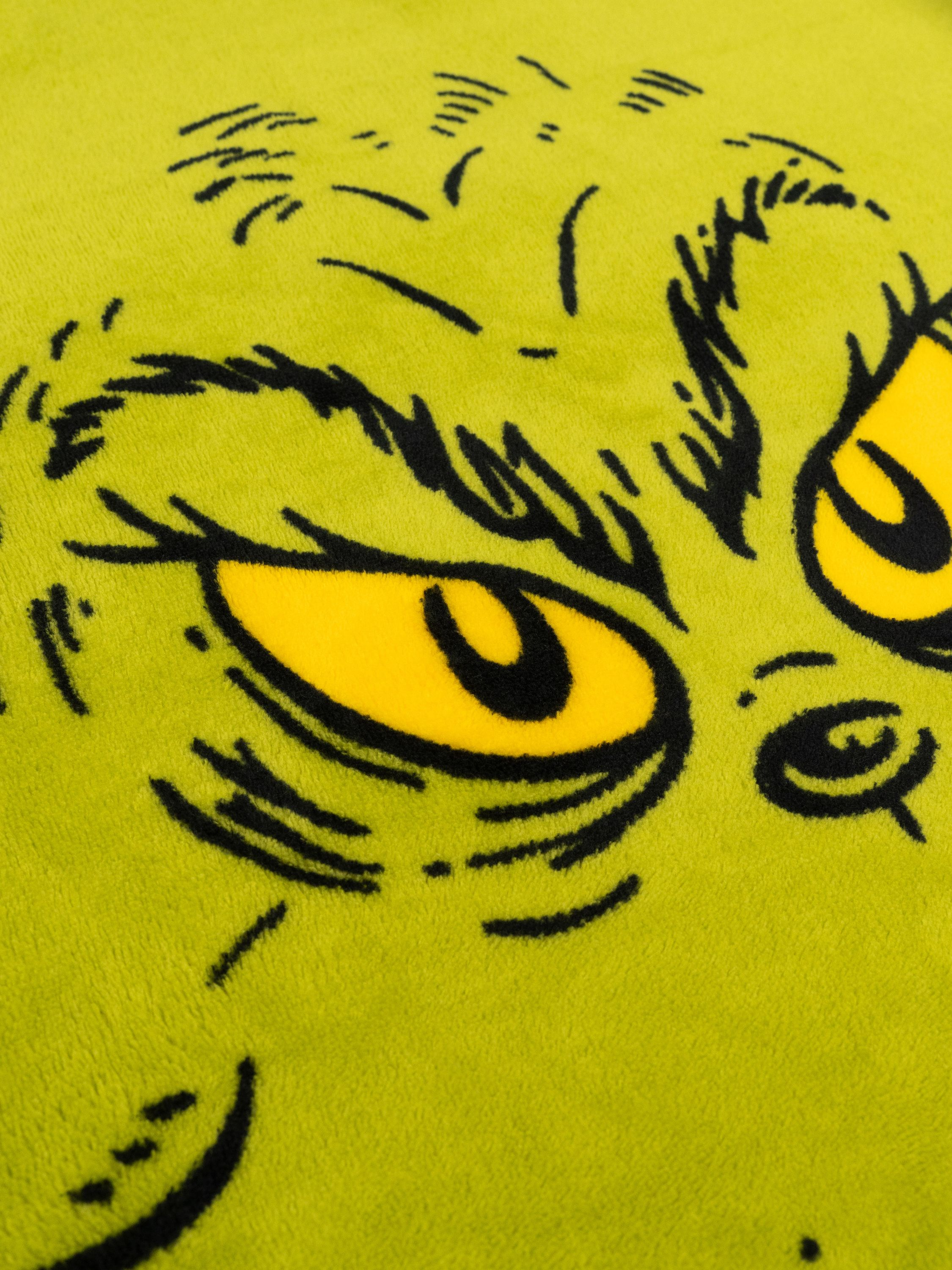 Nahaufnahme eines grünen Grinch-Gesichts mit gelben Augen und schwarzen Details. Das Bild hat einen weichen, strukturierten Hintergrund.