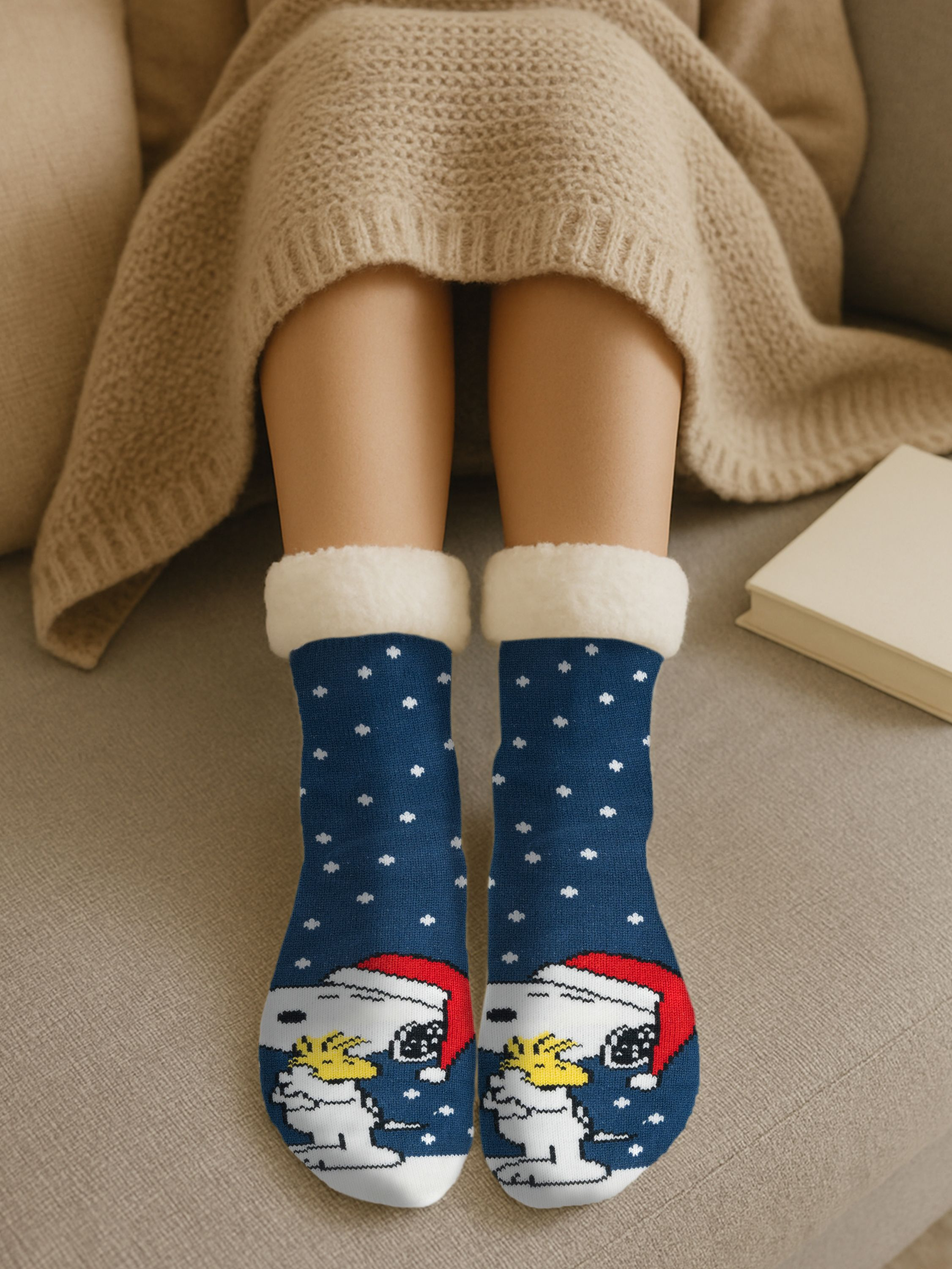 Person in blauen Snoopy-Socken mit weißen Punkten und weißem Besatz, trägt einen beigen Pullover, sitzt auf einem Sofa mit einem Buch.