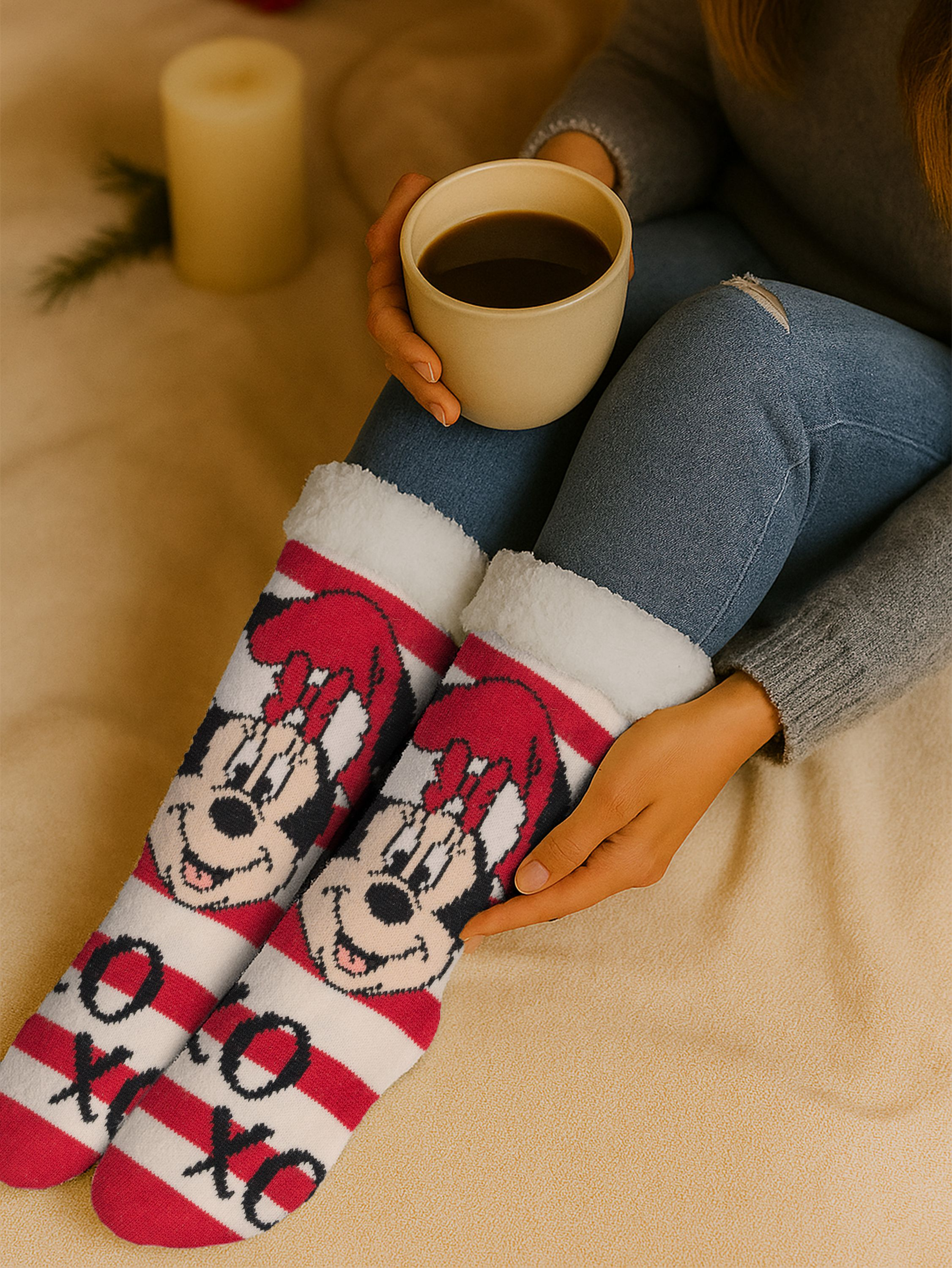 Person trägt Minnie-Mouse-Socken und hält eine Kaffeetasse. Eine Kerze und eine Decke sind im Hintergrund.
