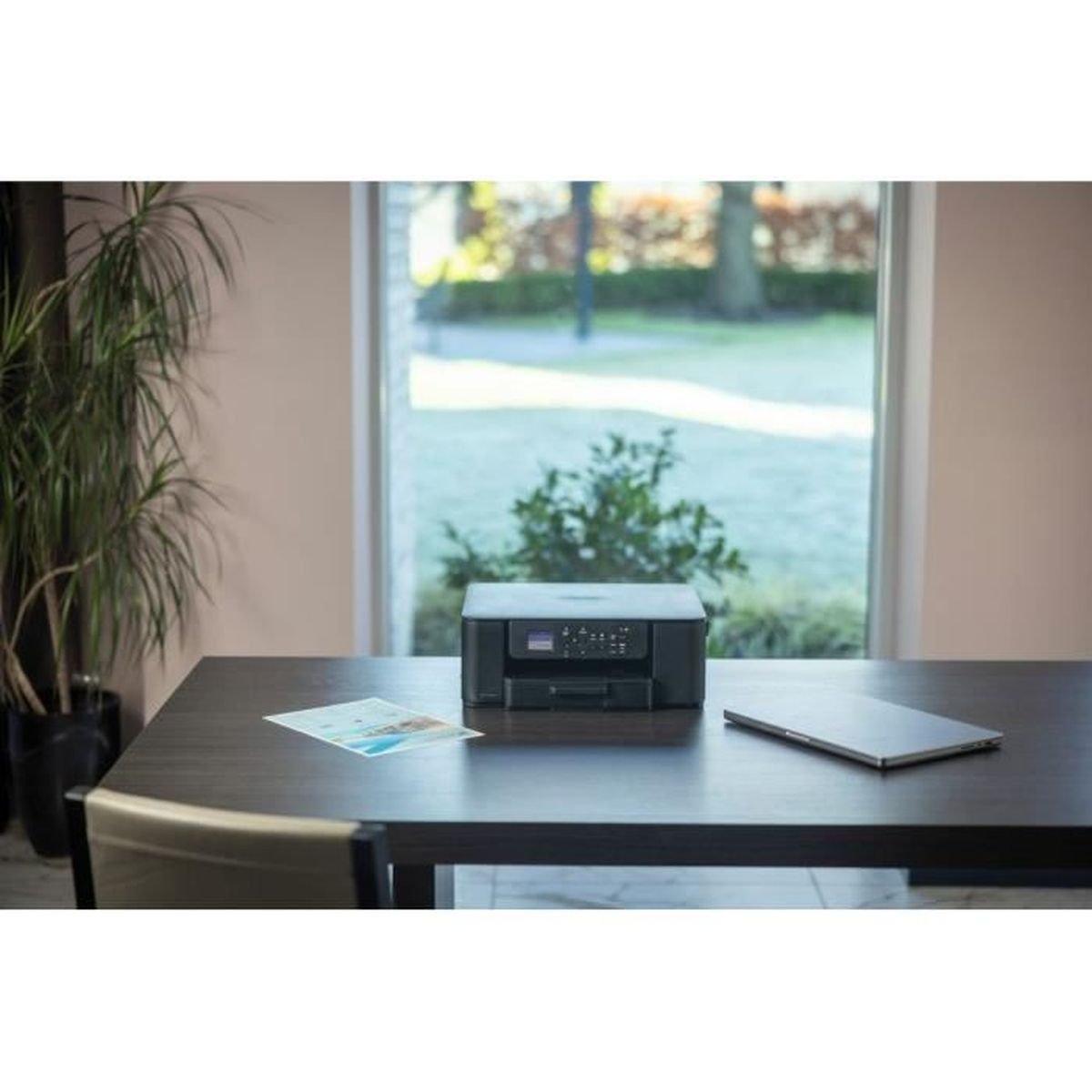 Czarna drukarka, laptop i dokument na ciemnym drewnianym biurku, z oknem w tle. Roślina po lewej stronie.