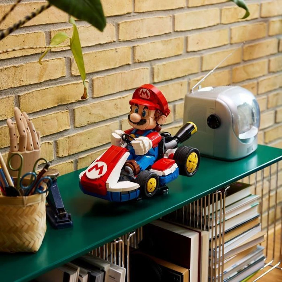Lego Mario w gokarcie na zielonej półce, z ceglanym murem i małym radiem. Obecne są również książki i materiały artystyczne.