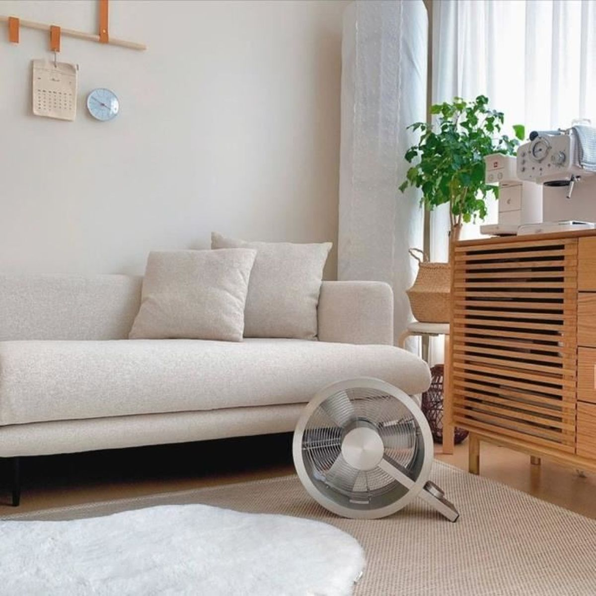 Scène de salon avec canapé, ventilateur et meuble en bois. Une horloge et un calendrier sont accrochés au mur. Un tapis blanc est au sol.
